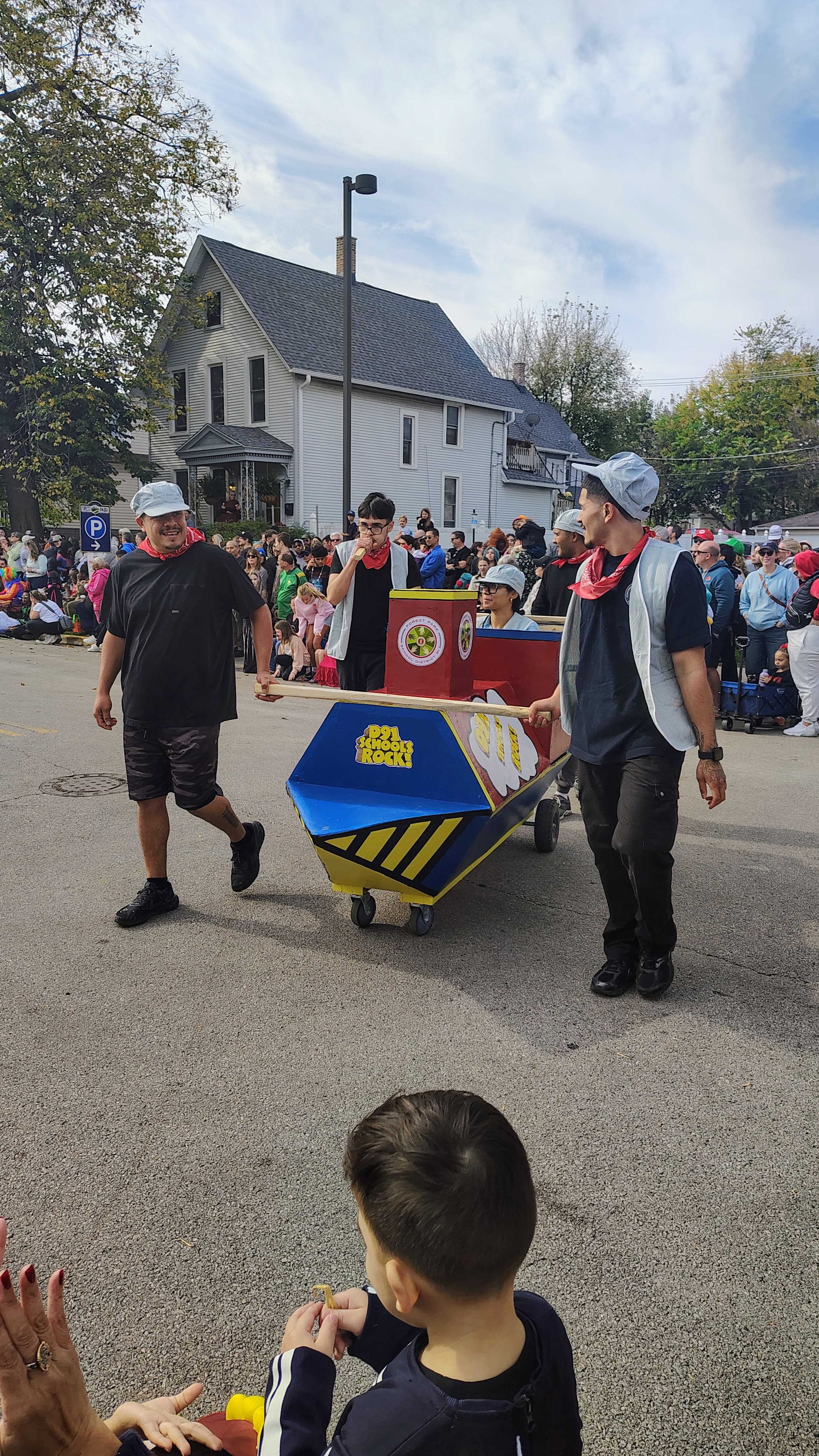A crew in red bandanas and caps pushing a train-shaped casket painted in red, blue, and yellow reading D91 Schools Rock