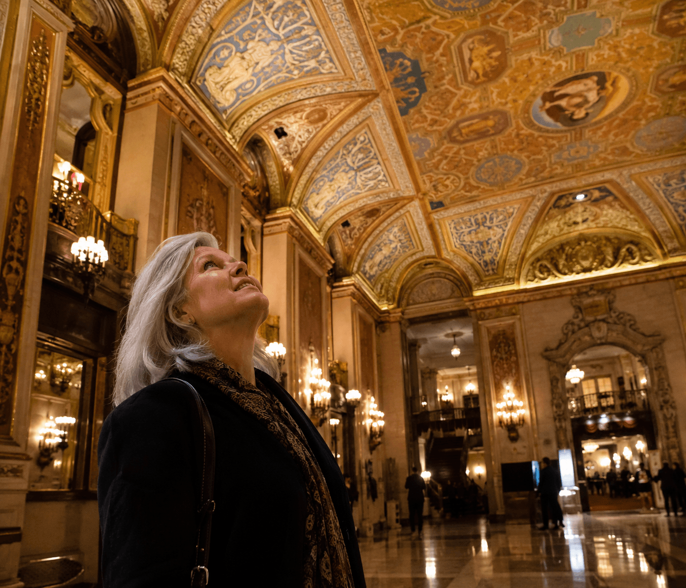 A silver-haired woman standing in a grand old theater lobby, looking up in awe at an ornate gilded ceiling