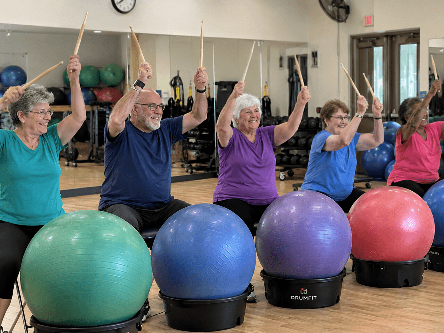 A row of seniors on exercise balls, drumsticks raised in sync, smiling mid-class