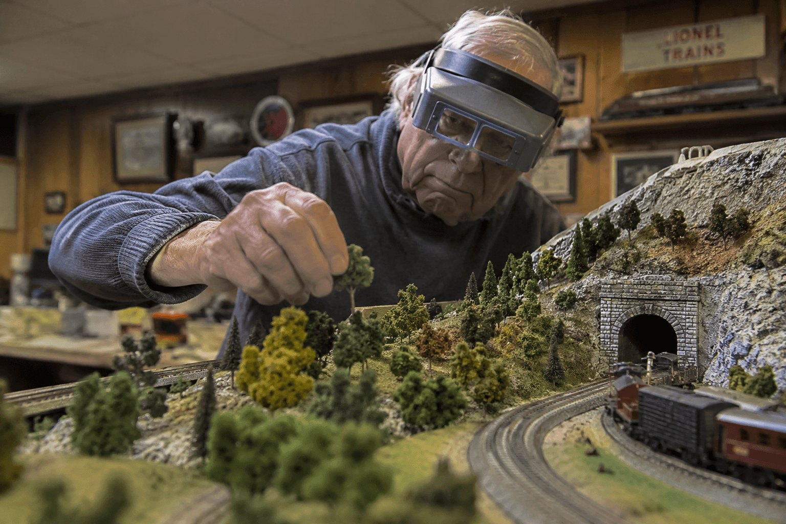 An older man with magnifying glasses carefully placing a tiny tree on an elaborate model railroad layout
